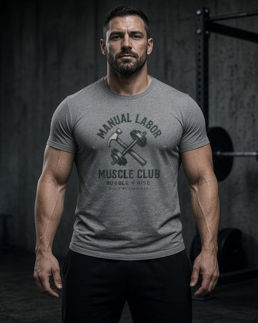 Man in a dark gym wearing the Rubble and Rise Manual Labor Muscle Club tee in Athletic Heather